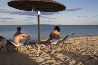 Tatil veya balayı Çift. Erkek ve kadın üstsüz deniz, arka görüş üzerinde manzarasının tadını çıkarın. Deniz Resort aşık çift şezlong kum plaj şemsiyesi altında oturur. Balayı konsepti.