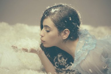 Yan görünümü esmer kız yatak odasında tüyler ile oynarken. Mavi ipek gecelik tüylü yatak, havada akan kabartmak yatan genç modelinde. Pijama partisi ve pijama partisi konsepti.