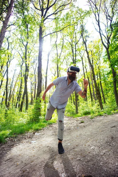 Bearded man having 3D experience in VR goggles. Excited bearded hipster testing new gadget in forest. Man with trendy beard and mustache running in virtual reality, digital era concept.