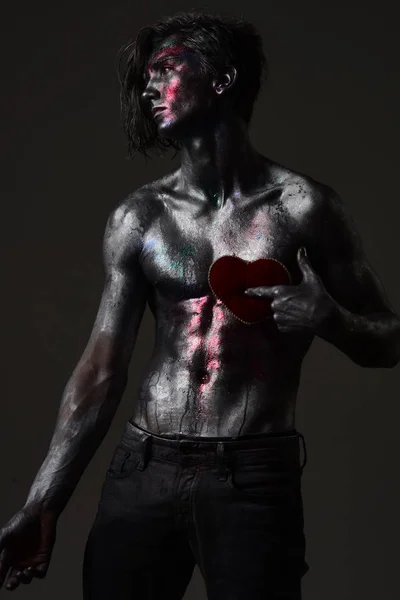 Man with nude torso holds red plush soft heart toy on chest, dark background. Loneliness and brake up concept. Guy on sad face with make up, covered with shimmering silver paint and colorful glitters.