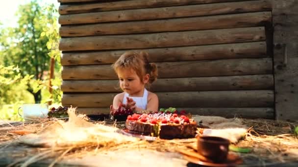 Schattige kleine jongen in witte schort eet cake van het fruit en kopje melk aan tafel buiten op houten — Stockvideo