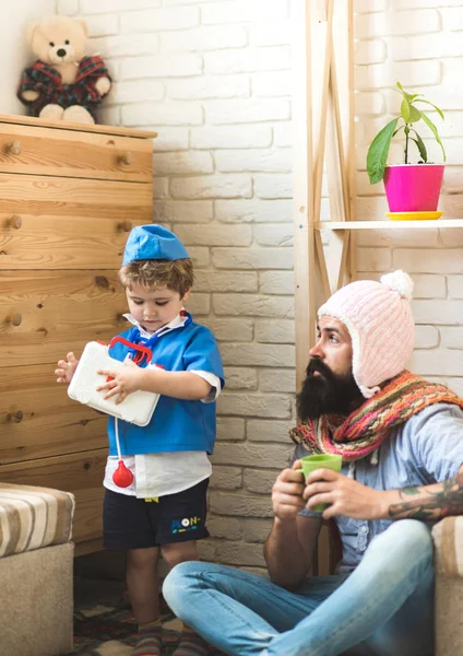 Boy with first aid kit over white brick wall. Father and son playing hospital at home, medicine concept. Bearded man pretending to be patient in game with kid.