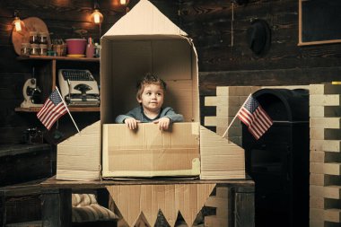 Kids playing with toys. Boy play with rocket, cosmonaut sit in usa rocket made out of cardboard box. Kid on calm face sit in cardboard hand made rocket with usa flags. Child boy play american clipart