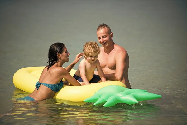 家族での休暇の概念。父とマットレスに近い母は、息子と一緒に泳ぐ。家族は、一緒に時間を過ごす楽しんでいます。エアマットレス パイナップル形の両親と海、海の上に座っているかわいい子少年 — ストック写真