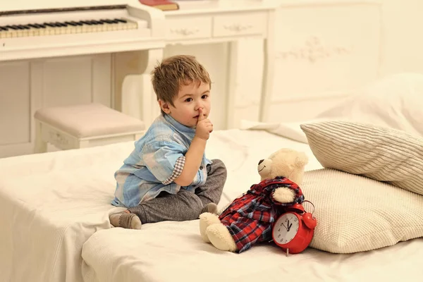 Barn som leker med leksaker. Barn i sovrum med tystnad gest. Kid sätta plysch Björn nära kuddar och väckarklocka, lyxig interiör bakgrund. Dags att sova koncept. Pojke med lugna ansikte sätter favoritleksak — Stockfoto