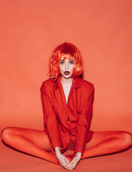 Excited young woman in fancy bob style red wig posing isolated on red background. Surprised girl in red outfit sitting on floor in butterfly stretch. Fitness, stretching and flexibility concept.