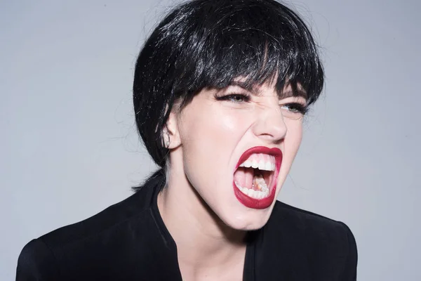 Woman with attractive red lips shouting, close up. Lady in black wig with make up screaming on grey background. Angry boss concept. Girl on scandalous shouting face wears formal jacket.