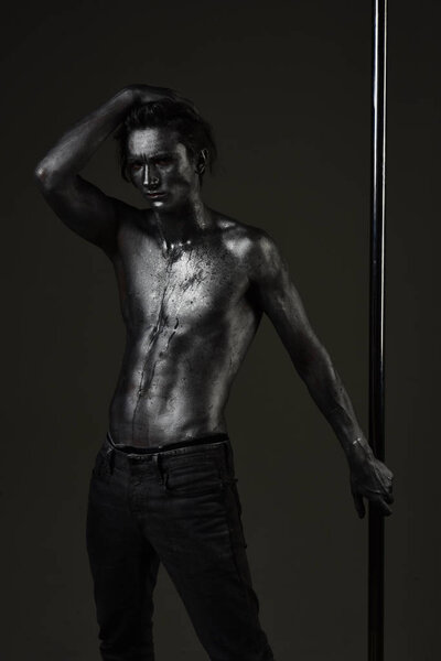 Macho with naked chest, athlete, sportsman hold metallic pole. Man with nude torso covered with shimmering silver paint, dark background. Fashion shot of young sexy mysterious athlete. Fashion concept