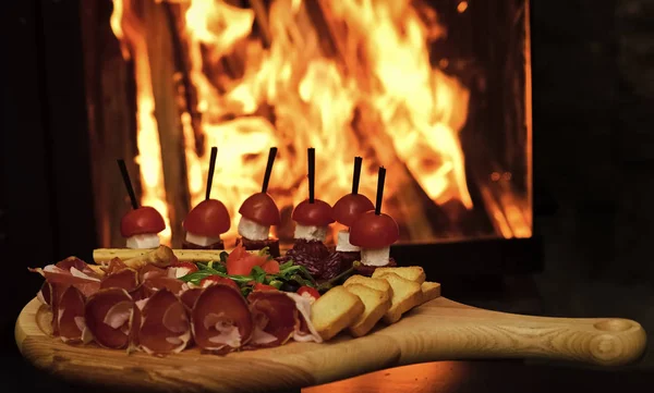 Junta con bocadillos en mesa de madera, fuego en el fondo. Aperitivos fríos con tomates, salchichas, salami, jamón, rúcula, aceitunas y pan. Snacks apetitosos servidos en mesa redonda. concepto de plato restaurante —  Fotos de Stock