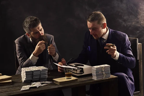 Un hombre elegante de traje. Hombres sentados a la mesa con montones de dinero y máquina de escribir. Empresa dedicada a negocios ilegales. Concepto de negocio ilegal. Empresarios discutiendo trato ilegal mientras beben y —  Fotos de Stock