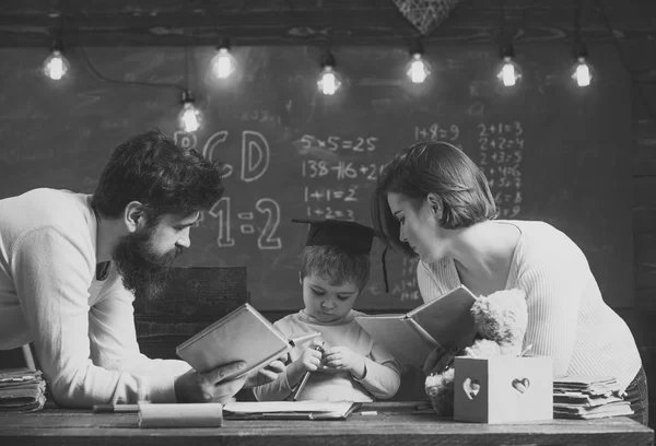 Niño jugar concepto de educación en casa. Padre y madre leyendo libros, enseñando a su hijo, pizarra en el fondo. La familia se preocupa por la educación de su hijo. Niño con gorra de graduado aburrido de estudiar . — Foto de Stock