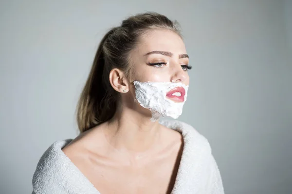 Side view woman with shaving foam on her face isolated on gray background. Girl with high ponytail wearing white bathrobe, transgender concept.