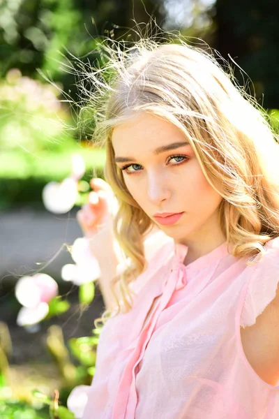 在自然背景下漂亮的金发女孩的肖像 年轻的女孩与卷曲的金色的锁穿着粉红色的衬衫摆在开花花园在阳光明媚的一天 — 图库照片