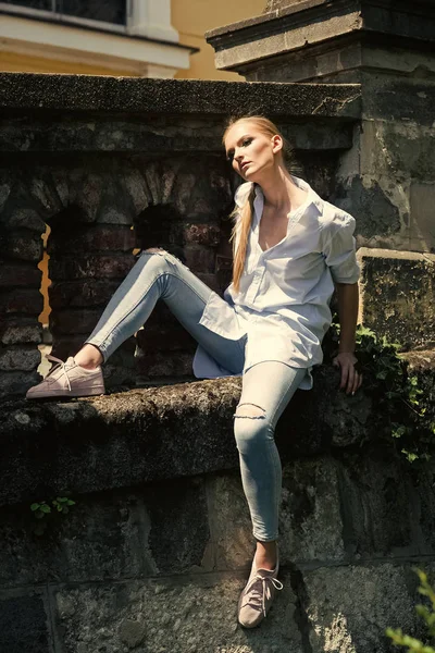 Fashion woman body. Woman in shirt and jeans sit on stone wall, fashion. Woman with long blond hair, hairstyle on sunny outdoor. Fashion model with makeup face, beauty. Girl style, beauty, look and