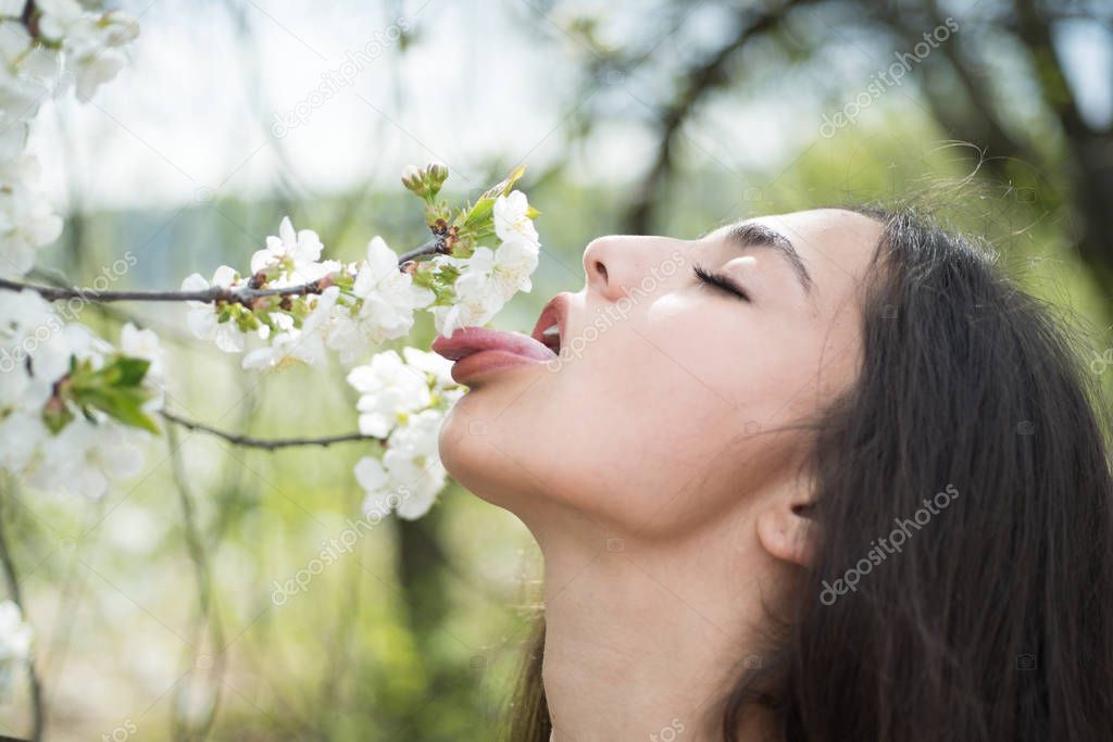 Sexy woman walking in blooming park or garden, spring concept. Young brunette with closed eyes licking white blossom. Side view portrait of beautiful girl with stick out tongue under flowering tree.