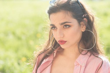 Genç esmer mükemmel makyaj, güneşli bir gün park veya bahçe keyfi moda güneş gözlüğü ile. Yeşil arka plan, doğal güzellik kavramı üzerinde pembe gömlekli büyük yuvarlak gümüş kulak ile çekici kız yüzük.