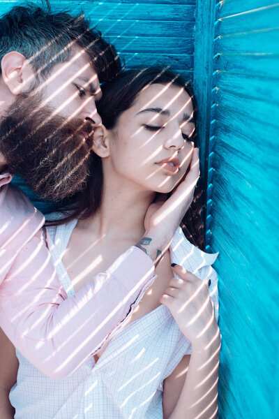Heat concept. Couple chilling near jalousie, stripes of sunlight and shadow on faces. Girl in unbuttoned shirt and man with beard. Couple in love on calm faces near turquoise jalousie.