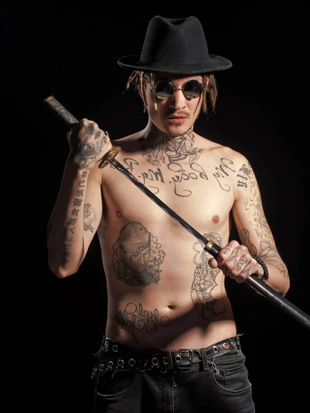 Pojke som drivs. Frågor inför pojkar. Mannen med nakna tatuerad torso i hatt hålla svärd Stockbild