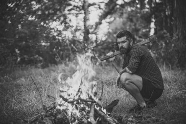 Adam ciddi duygu ile. Hipster adam balta ile doğal peyzaj üzerinde yaz gününde kamp ateşi ateşe bak. Seyahat, kamp, yürüyüş, tatil kavramı