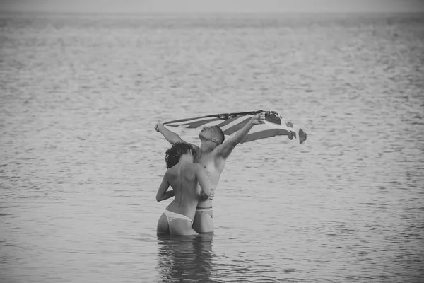 Una pareja sexy. Pareja enamorada de pie en el agua de mar, la superficie del mar en el fondo. Pareja en topless disfruta de vacaciones, el hombre sostiene la bandera de EE.UU. por encima de la cabeza. Concepto de libertad. Familia joven viajando de luna de miel — Foto de Stock