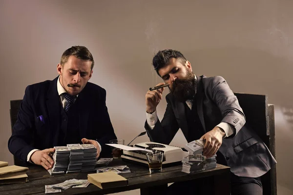 Man with serious emotion. Company engaged in illegal business. Businessmen discussing illegal deal while drinking and smoking, grey background. Men sitting at table with piles of money and typewriter