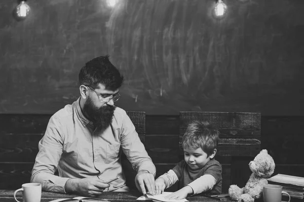 Chico feliz divirtiéndose. Profesor y niño pequeño sentados en el escritorio con libros y osito de peluche. Colegial jugando con copybook. Profesor estricto en gafas delante del tablero verde —  Fotos de Stock