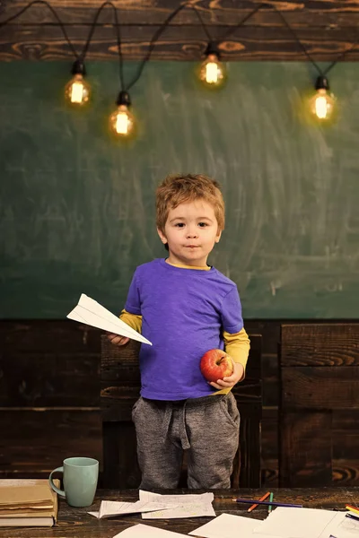 Kinderen spelen - gelukkig spel. Jongetje bedrijf papier vliegtuig en apple in zijn handen. Scholier in blauw T-shirt staande achter de tafel. Educatieve spelconcept — Stockfoto