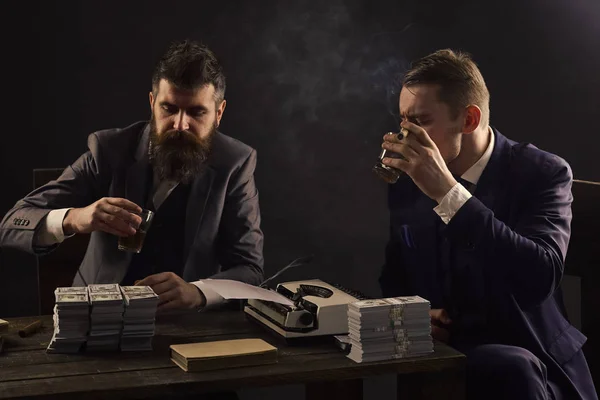 違法なビジネス コンセプトです。会社は、違法なビジネスに取り組んでいます。男性はお金とタイプライターの山でテーブルに座っています。飲酒や喫煙、暗い背景中違法契約を議論するビジネスマン. — ストック写真
