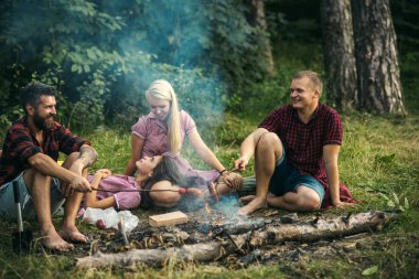 Neşeli grup ormanda piknik arkadaşıyla. Kamp ateşi etrafında konuşmaya gülümseyen, mutlu, Genç insanlar. Sosis pişirme oduncu gömlek erkeklerde.