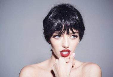 Kısa shinning siyah saçlı ve yana doğru seyir, flört ve iğfal yüzünü dokunaklı kırmızı dudaklar ile güzel kız. Kadın siyah peruk gri arka plan üzerinde izole çıplak makyajlı modelde.