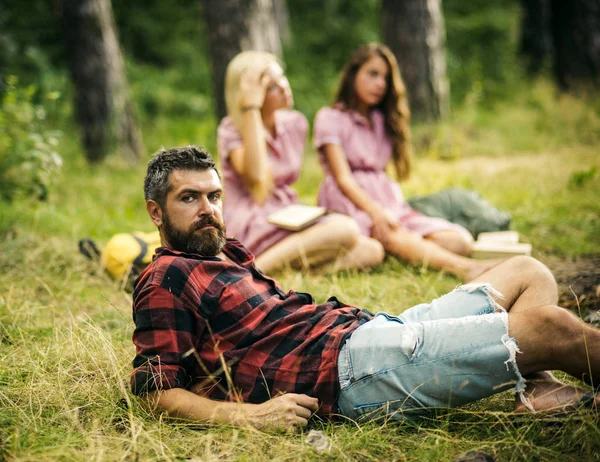Bearded man in lumberjack shirt spending time with friends outdoors. Handsome man lying on glass in woods. Outdoor recreation and nature concepts.