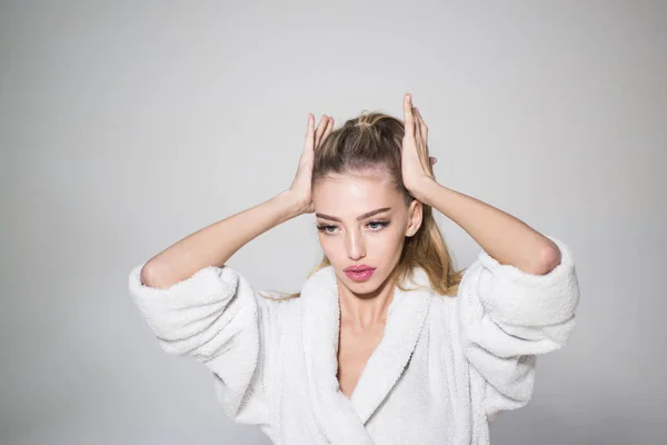 Young beautiful woman fixing her hair in bathroom. Attractive blond girl in white bathrobe isolated on gray background, morning make-up before work.