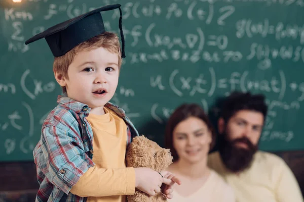 Support concept. Kid holds teddy bear and performing. Parents listening their son, chalkboard on background. Smart child in graduate cap like to perform. Boy presenting his knowledge to mom and dad.