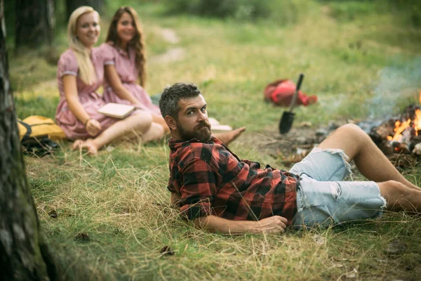 Bärtiger Mann im Holzfällerhemd, auf Gras liegend, Einheit mit der Natur. Gruppe junger Wanderer zeltet im Wald — Stockfoto