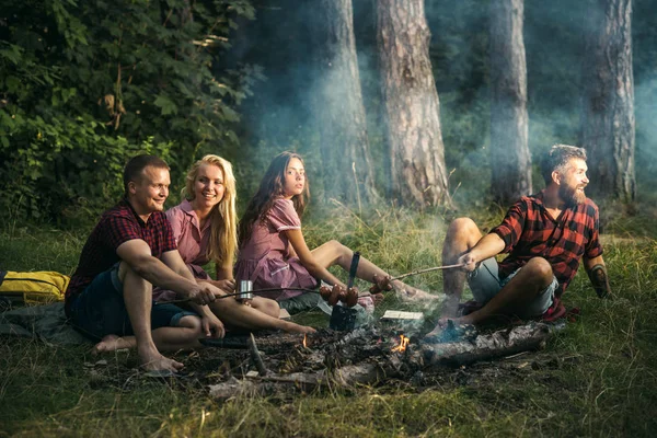 森の中でキャンプの若者のグループ。キャンプファイヤーでソーセージをフライパン 2 つのカップル。男を呼び出して、友達を歓迎を興奮させた。彼のガール フレンドは、マグカップからお茶を飲んでいる間に料理を男します。 — ストック写真