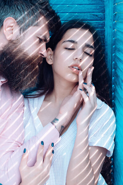 Couple chilling near jalousie, stripes of sunlight and shadow on faces. Heat concept. Couple in love on calm faces near turquoise jalousie. Girl in unbuttoned shirt and man with beard.