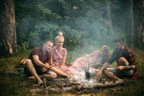 Две пары устраивают пикник в лесу. Бородач и его лучший друг готовят сосиски над огнем. Молодые туристы у костра вечером. Концепция дружбы и отдыха — стоковое фото