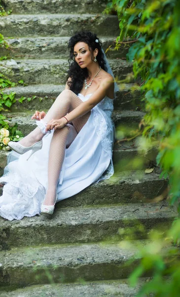 Girl with bridal makeup and hairstyle. Woman wear lace garter on leg. Sexy woman in stockings lingerie on wedding day. Bride in white dress sit on steps outdoor. Wedding fashion and accessory — Stock Photo, Image