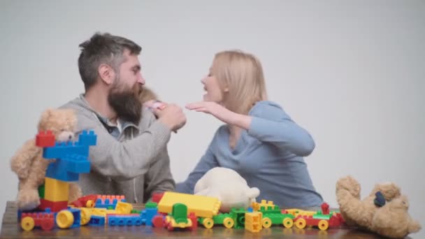 Joven juego familiar con bloques de plástico de construcción. Concepto de juegos familiares. Los padres y los niños con caras felices sostienen ladrillos rojos. Padre, madre e hijo en la sala de juegos sobre fondo de madera clara . — Vídeos de Stock