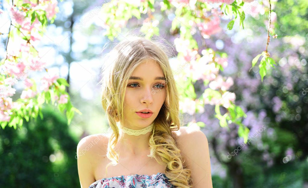 Portrait of blond lady with long curled hair and blue eyes in floral garden. Girl in vintage style outfit enjoying warm day in shade of blooming tree with tender pink flowers, spring time concept