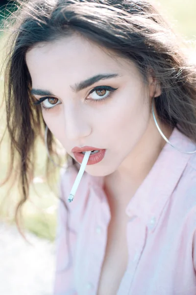 Mulher sensual em camisa rosa com maquiagem perfeita segurando cigarro na boca. Morena sexy com lábios vermelhos completos fumar, mau hábito, conceito de saúde — Fotografia de Stock