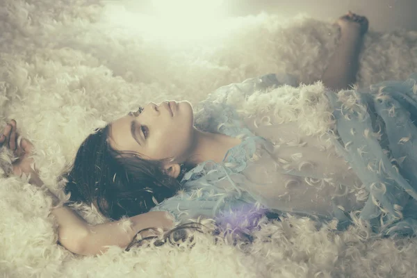 Мрійлива дівчина в блакитній нічній сукні лежить на м'якій купі білого пір'я. Красива молода жінка фантазує в ліжку, солодкі сни і концепція сну — стокове фото
