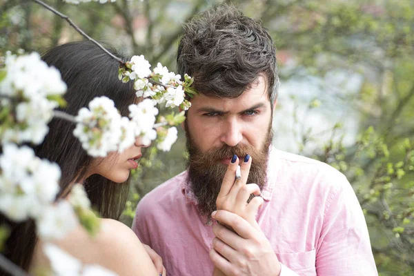 Bearded man in pink shirt smoking in flowery garden. Pretty female hidden behind branch of blooming tree holds cigarette, temptation concept