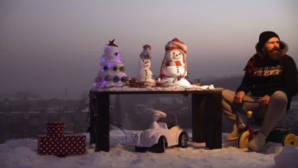 Snögubbe och snö xmas träd på vinterlandskap. Mannen i pilot glasögon och huva på cykel. Jul och nyår kul. Holiday fest koncept. Man rider trehjuling på grå himmel. — Stockvideo