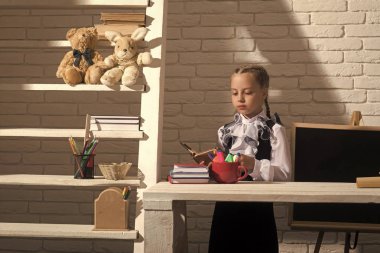 Eğitim ve okul kavramı dön. Beyaz tuğla zemin üzerine sınıfta çocuk. Ciddi bir yüz ile kız öğrenci yazı tahtası yakınındaki açık kitap tutar