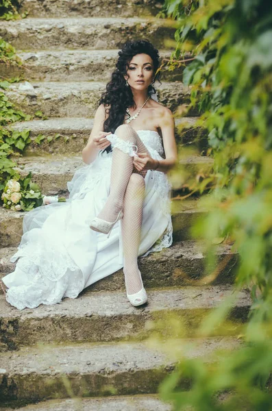 Bride in white dress sit on steps outdoor. Woman wear lace garter on leg. Sexy woman in stockings lingerie on wedding day. Girl with bridal makeup and hairstyle. Wedding fashion and accessory