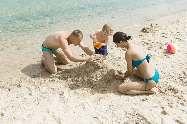 Viajar en familia con el niño el día de la madre o el padre. El amor y la confianza como valores familiares. Vacaciones de verano de familia feliz. Madre y padre con hijo hacen castillo de arena en la playa. Niño con padre y madre . — Foto de Stock