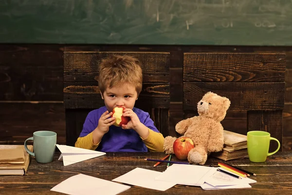 Cambio di scuola. Vacanza scolastica. Ragazzo affamato che morde mela in classe. Piccolo ragazzo che gioca con aereo di carta e orsacchiotto — Foto Stock