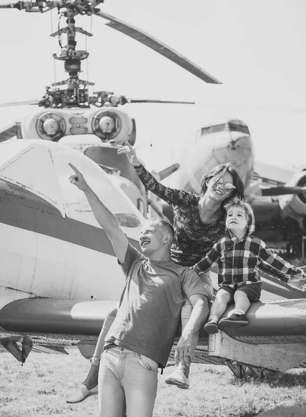 Family in the aviation museum. Mother, father and excited child sit on planes wing in aviation museum. Happy family looking on sky on excursion, helicopter or plane on background, sunny day — Stock Photo, Image