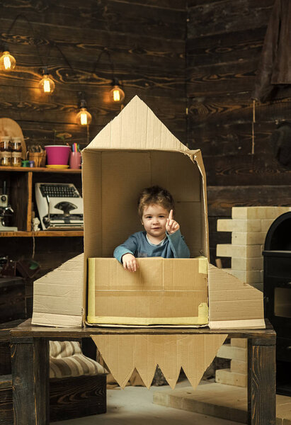 Childhood concept. Kid sit in cardboard hand made rocket, pointing upwards. Boy play at home with rocket, little cosmonaut sit in rocket made out of cardboard box. Child boy play cosmonaut, astronaut